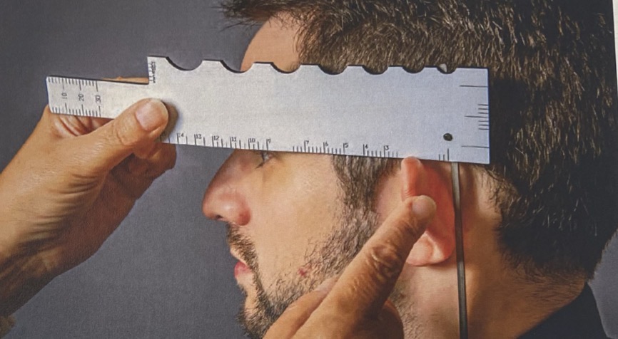 <p>Rotate either the short or long pointer so that it measures 90 degrees with the lower edge of the gauge.</p><p>View the patient's head from the side. Place the gauge with the 90 degree angle at the back of the ear and rest the gauge on the top of the ear and in line with the side of the frame.</p><p>Measure on the long scale to a position which just clears the lashes.</p><p>Ensure only one eye is used to judge this in order to prevent parallax issues.</p><p>The ruler should then be reversed to take measurements on the other side.</p>