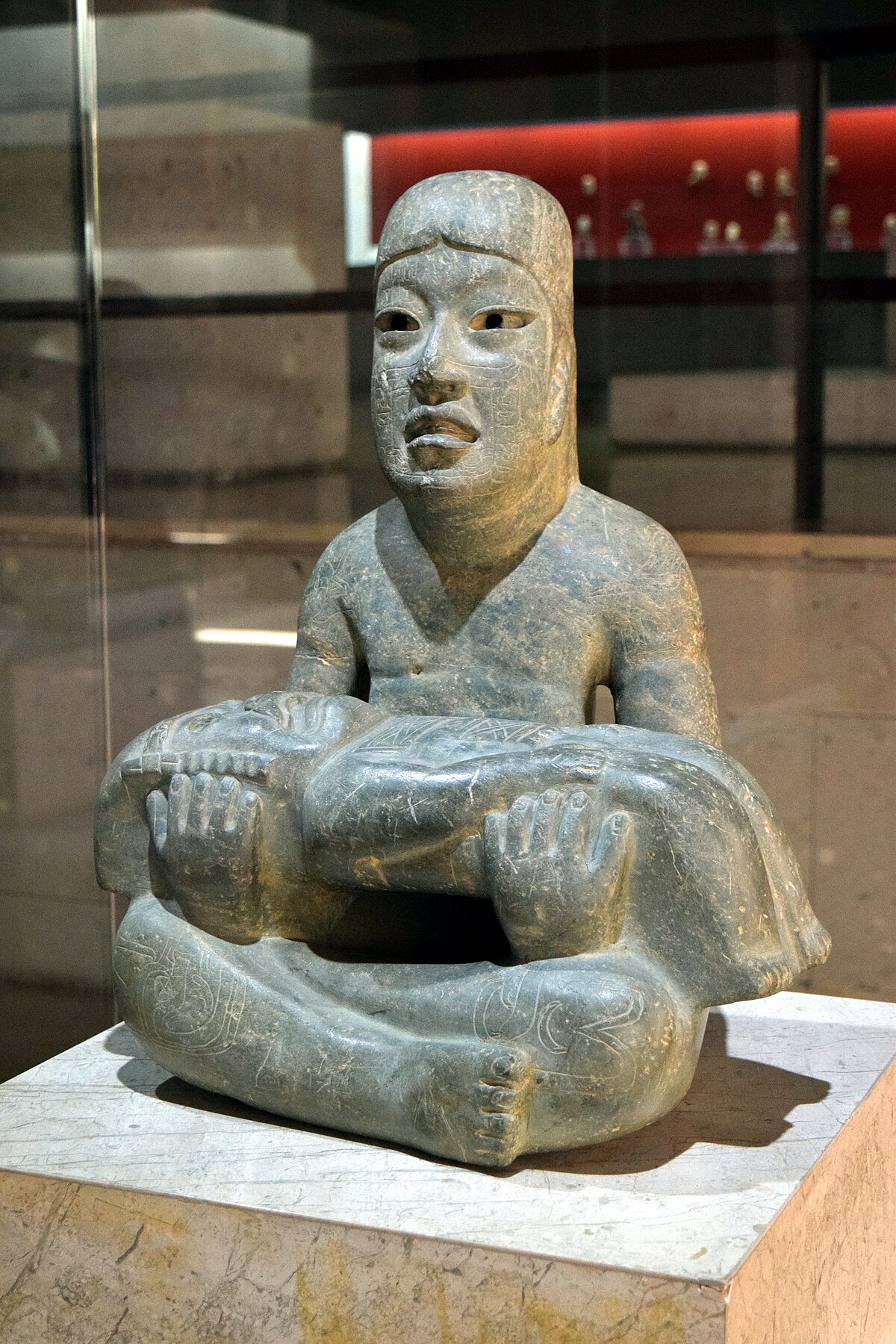 <p>Middle Formative. Olmec greenstone. Child holds baby in lap. Baby has face of Olmec rain deity. Knees, shoulders, face of child are incised with faces of other supernaturals, perhaps mapping out the cardinal directions.</p>