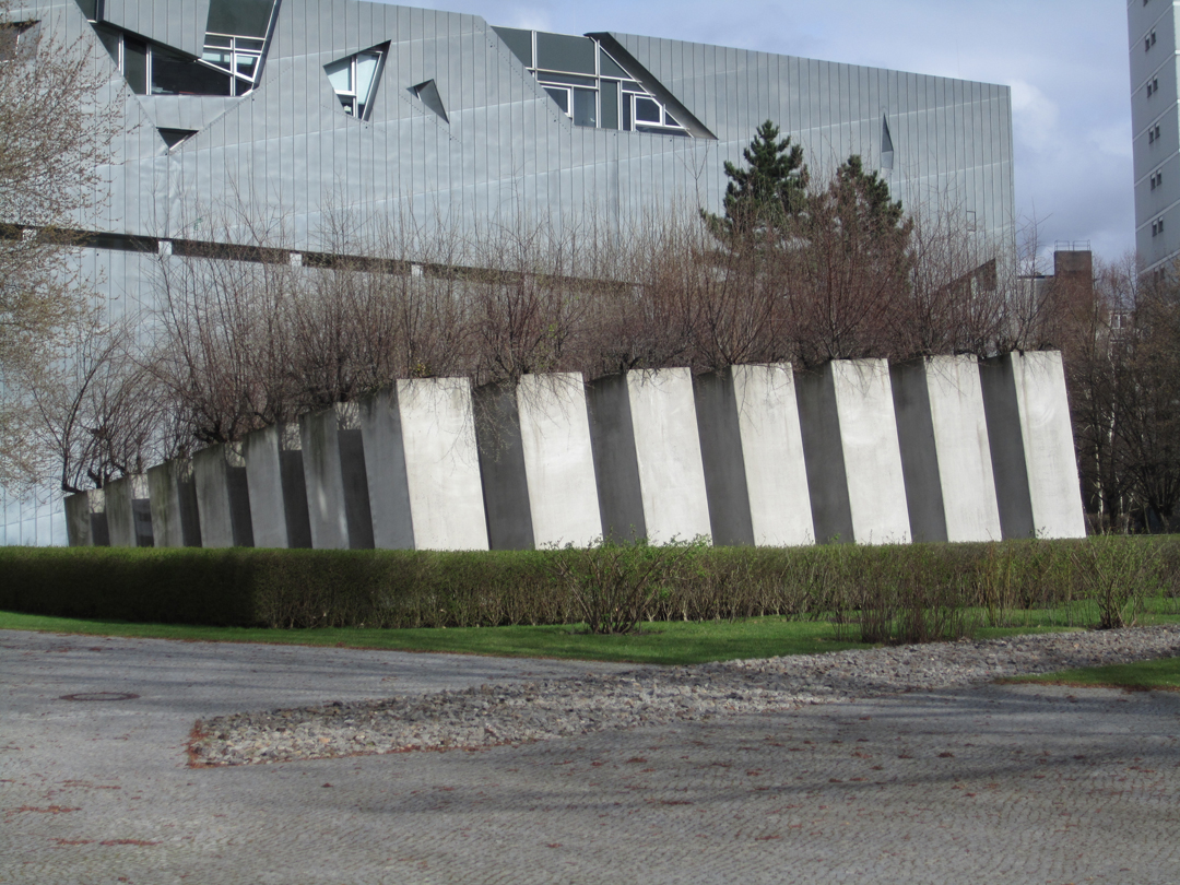 <ol><li><p><strong>49 concrete pillars</strong> - imposing, unevenly spaced columns represent instability and displacement of exile</p></li><li><p><strong>slanted ground</strong> - it disrupts the user’s sense of balance, creating a sense of dizziness and disorientation, mirroring the loss of stability faced by those in exile</p></li><li><p><strong>olive trees</strong> - these grow on top of the pillars, symbolising hope and peace</p></li><li><p><strong>axis of exile</strong> - the graden is accessed through an underground path - connecting it to the broader narrative of forced migration</p></li></ol><p></p>