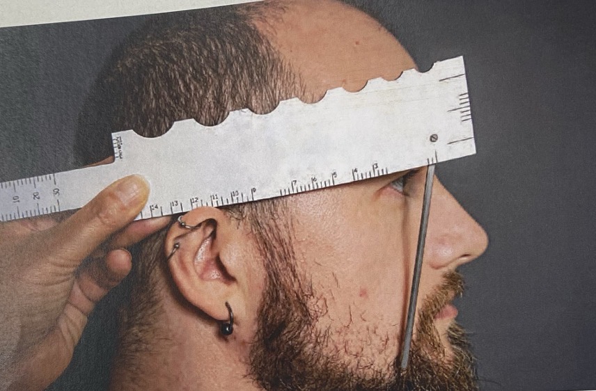 <p>View the patient's head from the right side. Place the gauge with the front surface forward and flat edge downwards resting on the top of the ear and in line with the side of the frame.</p><p>Rotate either the short or long pointer into a position representing the plane of the spectacle front and in a position which just clears the lashes, ensuring only one eye is used to judge this in order to prevent parallax issues.</p><p>The length to bend is measured on the long scale to the ear point and the angle of side is measured using the protractor scale.</p><p>The ruler should then be reversed to take measurements for the left side.</p>