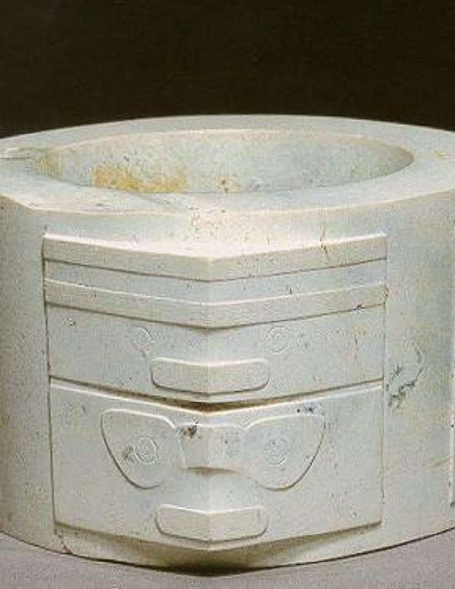 <p>A jade object with a circular hole within a square, featuring abstract designs and mask-like images.</p>